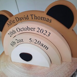 May include: A wooden bear-shaped keepsake with the name "Alfie David Thomas" and birth details: October 20th, 2023, 8lb 2oz, and 5:20am. The bear has tan, black, and white accents, and sits on a wooden base.