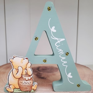 Pode incluir: Uma letra "A" de madeira pintada de verde claro, decorada com pequenas abelhas amarelas e pretas. A letra tem o nome "Aimee" escrito em branco. Uma figura do Ursinho Pooh est&aacute; ao lado da letra, perto de um pote de mel.
