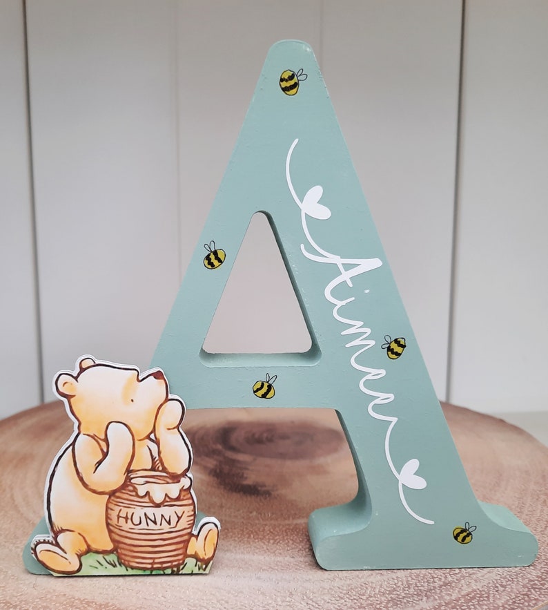 Pode incluir: Uma letra "A" de madeira pintada de verde claro, decorada com pequenas abelhas amarelas e pretas. A letra tem o nome "Aimee" escrito em branco. Uma figura do Ursinho Pooh est&aacute; ao lado da letra, perto de um pote de mel.