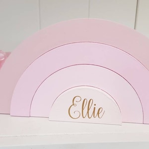 Pode incluir: Um arco-íris de madeira rosa com quatro arcos, o arco menor é branco com o nome "Ellie" escrito em letras douradas.