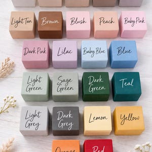 Pode incluir: Uma cole&ccedil;&atilde;o de vinte e quatro pequenos blocos de madeira quadrados, cada um pintado de uma cor diferente e rotulado com o nome da cor em escrita cursiva. As cores incluem branco, creme, bege e v&aacute;rias tonalidades de rosa, verde e cinza.