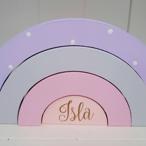 May include: A wooden rainbow stacking toy with three arches in shades of purple, gray, and pink. The smallest arch has the name "Isla" written in gold lettering.