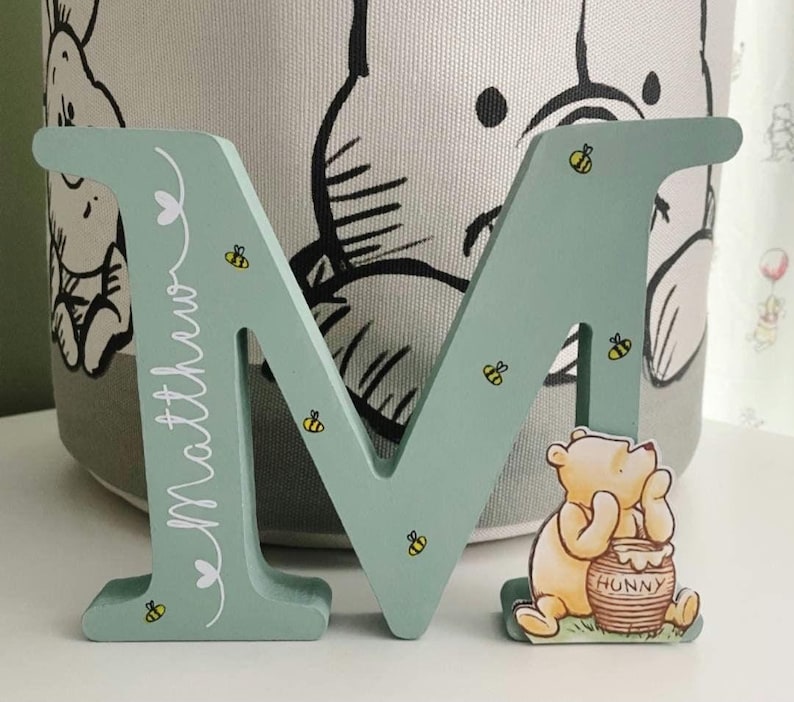 Pode incluir: Uma letra "M" de madeira verde-menta decorada com pequenas abelhas amarelas e pretas e o nome "Matthew" em escrita branca. Uma pequena figura do Ursinho Pooh est&aacute; sentada ao lado da letra, segurando um pote de mel.