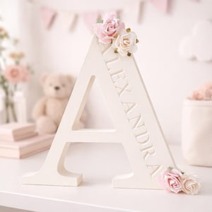 May include: A large, off-white wooden letter "A" with the name "ALEXANDRA" carved into the side. The letter is decorated with pink and cream-colored artificial roses. The background includes a teddy bear and decorative elements.