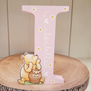 Pode incluir: Uma letra "I" de madeira rosa decorada com margaridas brancas e o nome "Isabella" em it&aacute;lico. Uma figura do Ursinho Pooh est&aacute; ao lado da letra, segurando um pote de mel com "Hunny" escrito. A letra est&aacute; em uma fatia de madeira.