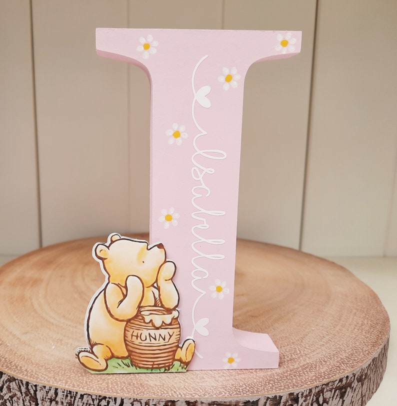 Pode incluir: Uma letra "I" de madeira rosa decorada com margaridas brancas e o nome "Isabella" em it&aacute;lico. Uma figura do Ursinho Pooh est&aacute; ao lado da letra, segurando um pote de mel com "Hunny" escrito. A letra est&aacute; em uma fatia de madeira.