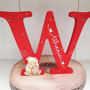 Pode incluir: Uma grande letra "W" de madeira vermelha com o nome "Winnie" em escrita branca, decorada com pequenas abelhas amarelas e pretas. Uma figura de Winnie the Pooh com um pote de mel &eacute; colocada em frente &agrave; letra. A letra est&aacute; em uma fatia de madeira.