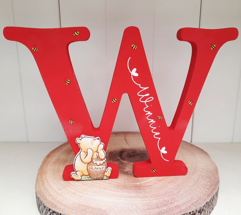 Pode incluir: Uma grande letra "W" de madeira vermelha com o nome "Winnie" em escrita branca, decorada com pequenas abelhas amarelas e pretas. Uma figura de Winnie the Pooh com um pote de mel &eacute; colocada em frente &agrave; letra. A letra est&aacute; em uma fatia de madeira.