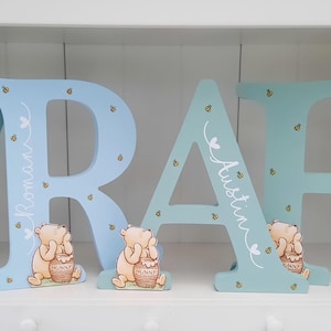 Pode incluir: Letras de madeira em azul claro e azul, soletrando nomes. Cada letra apresenta um personagem de Winnie the Pooh com um pote de mel. As letras s&atilde;o decoradas com pequenas abelhas e nomes em escrita cursiva branca.
