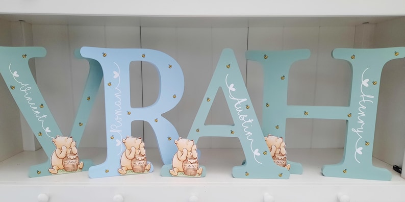 Pode incluir: Letras de madeira em azul claro e azul, soletrando nomes. Cada letra apresenta um personagem de Winnie the Pooh com um pote de mel. As letras s&atilde;o decoradas com pequenas abelhas e nomes em escrita cursiva branca.