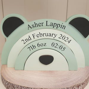 Puede incluir: Un apilador de madera en forma de oso verde menta con orejas negras y una nariz negra. El apilador muestra el nombre "Asher Lappin", la fecha de nacimiento, el peso (3,2 kg) y la hora (02:05). Descansa sobre una base de rodaja de madera.