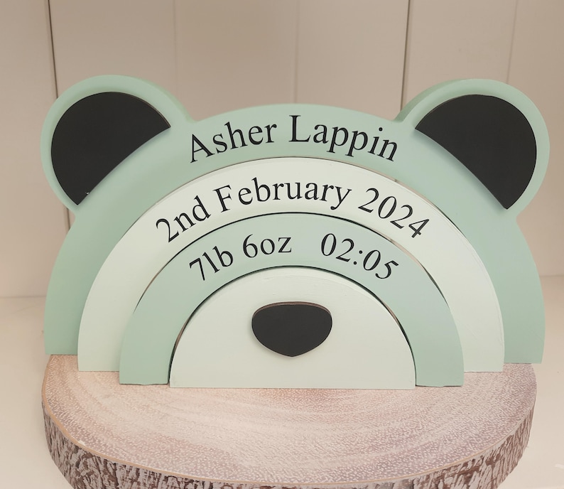 May include: A mint green bear-shaped wooden stacker with black ears and a black nose. The stacker displays the name "Asher Lappin", birthdate, weight (7lb 6oz), and time (02:05). It rests on a wooden slice base.