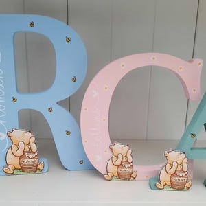 Pode incluir: Letras de madeira "RCA" em azul claro, rosa e verde-&aacute;gua. Cada letra apresenta um personagem de Winnie the Pooh com um pote de mel. As letras s&atilde;o decoradas com abelhas e os nomes "Roman", "Collins" e "Arlo".