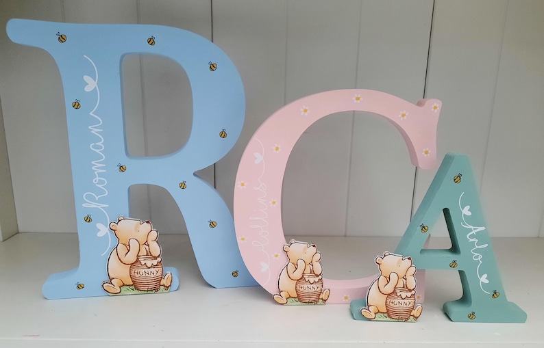 Pode incluir: Letras de madeira "RCA" em azul claro, rosa e verde-&aacute;gua. Cada letra apresenta um personagem de Winnie the Pooh com um pote de mel. As letras s&atilde;o decoradas com abelhas e os nomes "Roman", "Collins" e "Arlo".