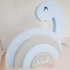 May include: A light blue wooden dinosaur with a grey and white rainbow stacked on top. The rainbow has white polka dots on the top layer. The bottom layer of the rainbow has the name "Noah" written on it.