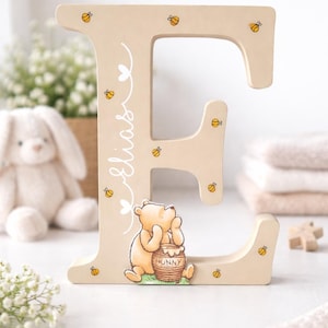 Pode incluir: Letra "E" de madeira bege claro, decorada com abelhas amarelas e uma ilustra&ccedil;&atilde;o de Winnie the Pooh. O nome "Elias" est&aacute; escrito em it&aacute;lico. Um charmoso elemento de decora&ccedil;&atilde;o infantil.