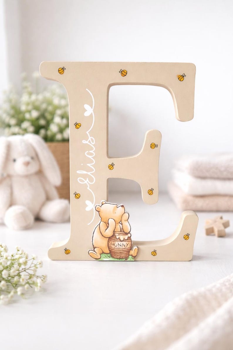 Pode incluir: Letra "E" de madeira bege claro, decorada com abelhas amarelas e uma ilustra&ccedil;&atilde;o de Winnie the Pooh. O nome "Elias" est&aacute; escrito em it&aacute;lico. Um charmoso elemento de decora&ccedil;&atilde;o infantil.