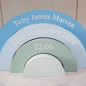 Puede incluir: Un arcoíris decorativo de madera en tonos azules y verdes. El arco superior dice "Toby James Marven". El segundo arco dice "22nd December 2024". El tercer arco muestra "22:06" y el arco inferior "7lb."