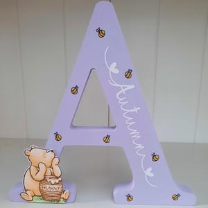 Pode incluir: Uma letra "A" de madeira lil&aacute;s com o nome "Autumn" em escrita branca, decorada com abelhas e cora&ccedil;&otilde;es amarelos e pretos. Um personagem de Winnie the Pooh com um pote de mel est&aacute; preso &agrave; base. Um item decorativo.