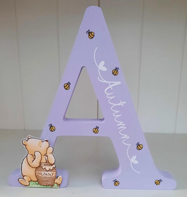 Pode incluir: Uma letra "A" de madeira lil&aacute;s com o nome "Autumn" em escrita branca, decorada com abelhas e cora&ccedil;&otilde;es amarelos e pretos. Um personagem de Winnie the Pooh com um pote de mel est&aacute; preso &agrave; base. Um item decorativo.