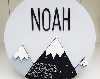 May include: A round, gray wooden sign with the name "NOAH" in black, raised letters. Below, a mountain scene features white-topped peaks, a geometric bear, and a starry night design. The sign is a decorative item.