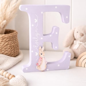Pode incluir: Uma letra "E" de madeira lavanda com bolinhas brancas e o nome "Evelyn" em itálico. Ilustração em aquarela de um coelhinho com casaco rosa. Decoração de quarto infantil.
