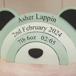 May include: A wooden bear-shaped decorative piece in pastel green, white, and black. The name "Asher Lappin" and birth details, including the date, weight (7lb 6oz), and time (02:05), are printed on the bear's body. The piece is displayed on a wooden surface.