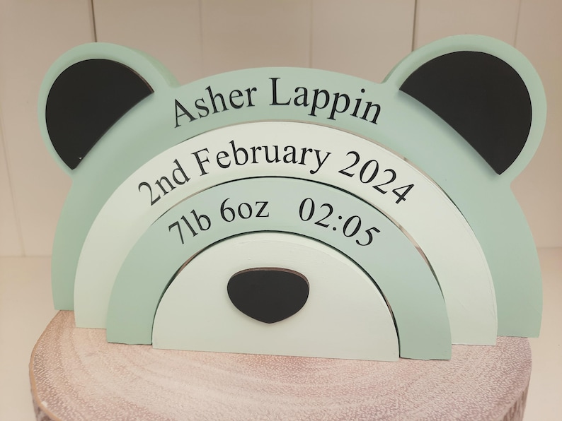 May include: A wooden bear-shaped decorative piece in pastel green, white, and black. The name "Asher Lappin" and birth details, including the date, weight (7lb 6oz), and time (02:05), are printed on the bear's body. The piece is displayed on a wooden surface.