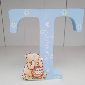 Pode incluir: Uma letra "T" de madeira azul clara decorada com margaridas brancas e o nome "Tommy" em escrita cursiva. Um personagem de Winnie the Pooh segura um pote de mel. O item &eacute; decorativo.