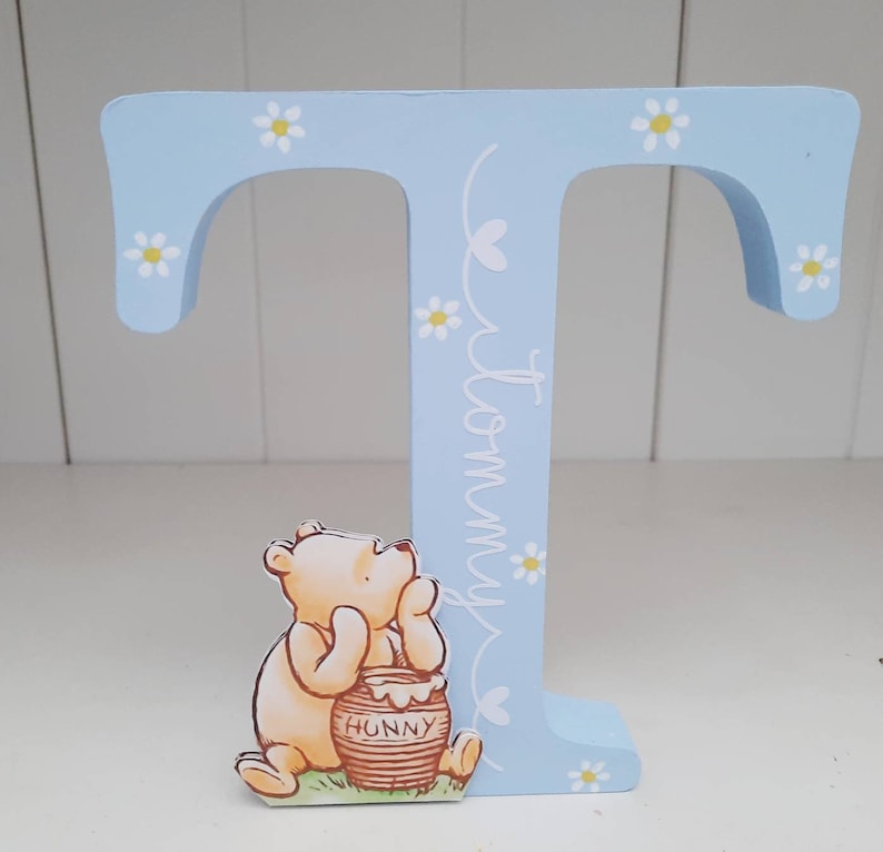 Pode incluir: Uma letra "T" de madeira azul clara decorada com margaridas brancas e o nome "Tommy" em escrita cursiva. Um personagem de Winnie the Pooh segura um pote de mel. O item &eacute; decorativo.