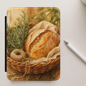 May include: A rectangular e-reader cover featuring a printed illustration of a round loaf of crusty bread inside a woven wicker basket, accompanied by a sprig of green rosemary and a small ball of twine resting on a light-colored fabric liner.