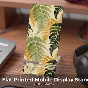 Puede incluir: Un soporte rectangular para móvil con fondo color crema y un patrón repetitivo de hojas de helecho estilo acuarela en tonos verde oscuro, verde oliva y amarillo dorado. El soporte cuenta con una base y un apoyo de acrílico transparente.