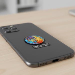 Puede incluir: Un smartphone negro con una pegatina redonda y colorida con un diseño de fuego y agua. La pegatina tiene el texto "EMF SHIELD PROTECTION & HARMONY". El teléfono está sobre una superficie de madera clara.