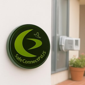 May include: A circular, dark green sign displaying the text "Safe Connect P:US" in a curved font. The sign features a green design of a curved shape and a bird. It is mounted on a white wall, near a window.