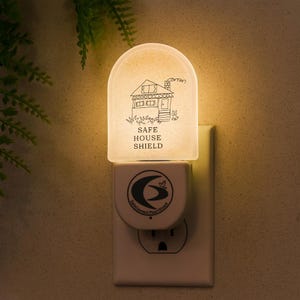 May include: A white night light plugged into a wall socket. The night light features a house illustration and the text "SAFE HOUSE SHIELD". The lower section displays a logo with the words "SafeConnect Plus Shield".