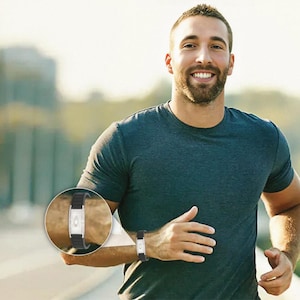 May include: A black and silver fitness tracker worn on the wrist. The tracker has a rectangular silver face with a small screen and is attached to a black band. The person is wearing a dark gray t-shirt.