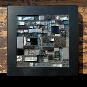 May include: Square mosaic art piece with a black wooden frame. The mosaic is composed of various shapes and sizes of glass tiles in shades of black, silver, and teal. The tiles have a reflective, textured surface.