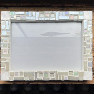 May include: A rectangular picture frame featuring a mosaic border composed of various white, off-white, and iridescent glass tiles. The frame surrounds a plain white rectangular center, with the mosaic pieces arranged in a varied, non-uniform pattern.
