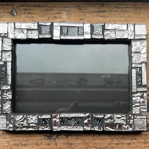 May include: Rectangular silver mosaic picture frame. The frame is made of small, irregularly shaped pieces of silver metal, creating a textured surface. The frame surrounds a dark, reflective surface, likely glass. The frame is set against a wooden background.