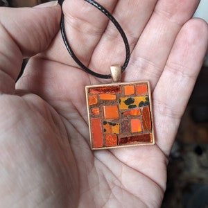 May include: Square pendant with a mosaic design in shades of orange, brown, and yellow. The pendant is set in a copper-colored frame and hangs from a black cord. The mosaic pattern features various shapes and sizes of tiles.