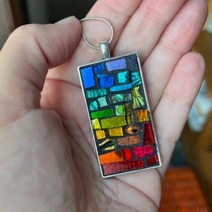 May include: A rectangular pendant with a mosaic design featuring a spectrum of colors, including blue, green, yellow, and red. The pendant is set in a silver-colored frame and hangs from a silver chain. The mosaic is made of small, irregular pieces.
