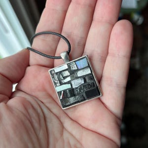 May include: Square pendant necklace with a mosaic design. The pendant features a silver-colored frame and a mosaic of black, white, and iridescent glass pieces. The necklace has a black cord.