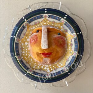 May include: A decorative wall plate featuring a central three-dimensional yellow sun face with red cheeks, a prominent nose, and red lips. The sun is mounted on a circular plate with a blue border, floral patterns, and white dots, set against a clear glass base.
