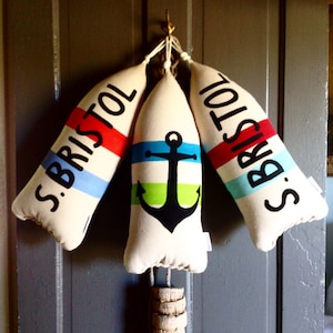 May include: Three white fabric buoy-shaped ornaments with red, blue, and green stripes. The center ornament has a black anchor with a green and blue stripe. Each ornament has the text "S. Bristol" in black.