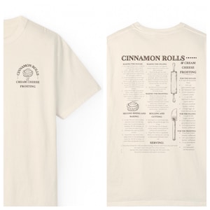 Puede incluir: Una camiseta beige con una receta de rollos de canela impresa en la espalda. La receta incluye instrucciones para hacer la masa, el relleno, el glaseado y el servicio. La parte delantera de la camiseta tiene un gráfico simple de un rollo de canela con el texto "Cinnamon Rolls & Cream Cheese Frosting".