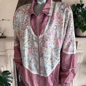 Könnte beinhalten: Vintage-Jacke mit Reißverschluss und floralem Muster in Rosa, Weiß und Blau. Die Jacke hat einen hohen Kragen und eine einfarbige rosa Blende auf der Vorderseite und an den Ärmeln. Die Jacke wird auf einer Schaufensterpuppe präsentiert.