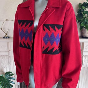 May include: A red wool jacket with a black and purple geometric design. The jacket has a full zipper, a collar, and ribbed cuffs and hem. The jacket is displayed on a mannequin.