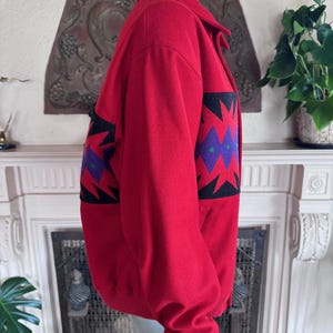 May include: A red wool jacket with a black, purple, and green geometric pattern on the sleeves. The jacket has a collared neckline, a zipper closure, and ribbed cuffs. The jacket is displayed on a mannequin.