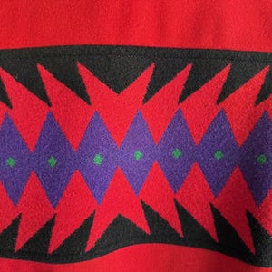May include: Close-up of a red fabric with a black, red, purple, and green geometric pattern. The pattern features red triangles and purple diamonds with green dots, set against a black background.