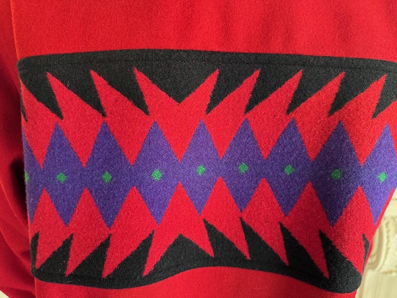 May include: Close-up of a red fabric with a black, red, purple, and green geometric pattern. The pattern features red triangles and purple diamonds with green dots, set against a black background.